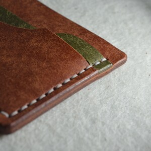 May include: Close-up of a brown leather wallet with olive green accents. The wallet features white stitching along the edges and a textured surface. The design includes card slots and a minimalist aesthetic.