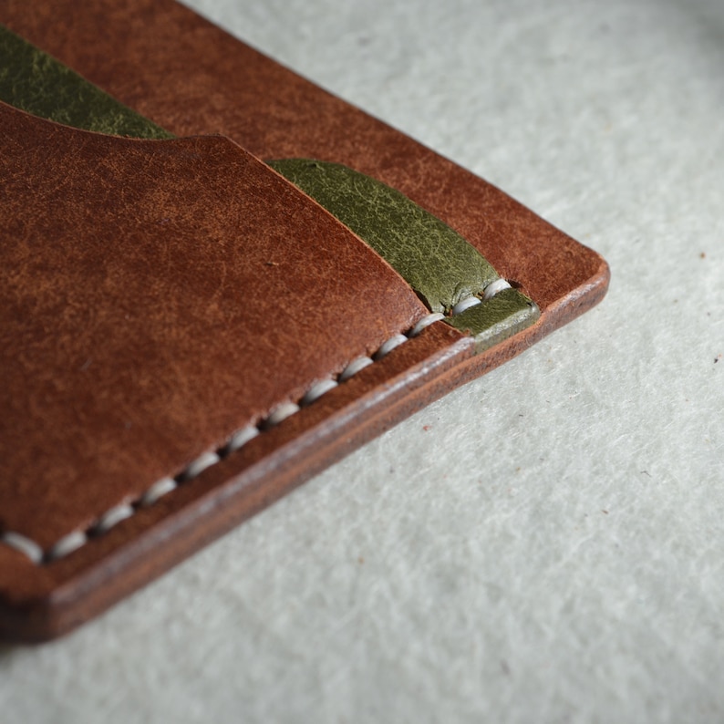May include: Close-up of a brown leather wallet with olive green accents. The wallet features white stitching along the edges and a textured surface. The design includes card slots and a minimalist aesthetic.