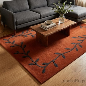May include: A rectangular area rug in a deep burnt orange color featuring a dark gray vine and leaf pattern that borders the perimeter. The rug is made of a textured material and is positioned on a light-colored wooden floor beneath a coffee table.
