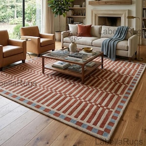 May include: A rectangular area rug featuring a geometric pattern of alternating terracotta orange and cream-coloured bars, bordered by a repeating sequence of small blue and cream squares, laid out on a wooden floor in a living room setting.