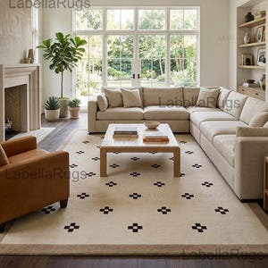Puede incluir: Una alfombra rectangular grande con una base de color crema y un patrón repetitivo de pequeños motivos en forma de cruz de color marrón oscuro. La alfombra está colocada en una sala de estar bajo una mesa de centro clara, con el texto LabellaRugs visible.
