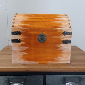 May include: A wooden treasure chest with a hinged lid and a metal latch. The chest is painted a light orange color and has a distressed finish. The chest is sitting on a wooden table.