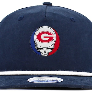 May include: A navy blue baseball cap with a white rope trim around the brim. The front of the cap features a red, white, and blue circular patch with a white "G" inside a red oval. The patch is a combination of the University of Georgia logo and the Grateful Dead's "Steal Your Face" logo.