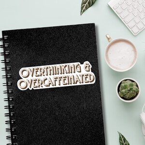 May include: A black notebook with a white sticker that says "Overthinking & Overcaffeinated" in brown text.