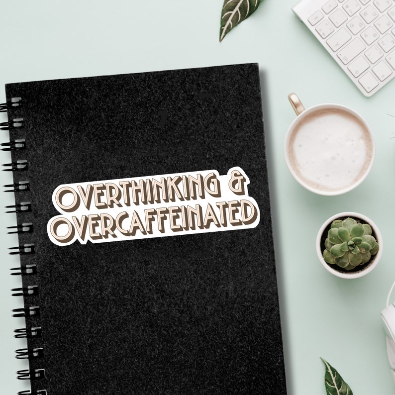May include: A black notebook with a white sticker that says "Overthinking & Overcaffeinated" in brown text.