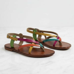 Puede incluir: Un par de sandalias de cuero con suela marrón y correas coloridas. Las correas son verdes, amarillas, naranjas, rosas y turquesas. La correa del tobillo tiene una hebilla. Las sandalias están sobre una superficie de mármol blanco.
