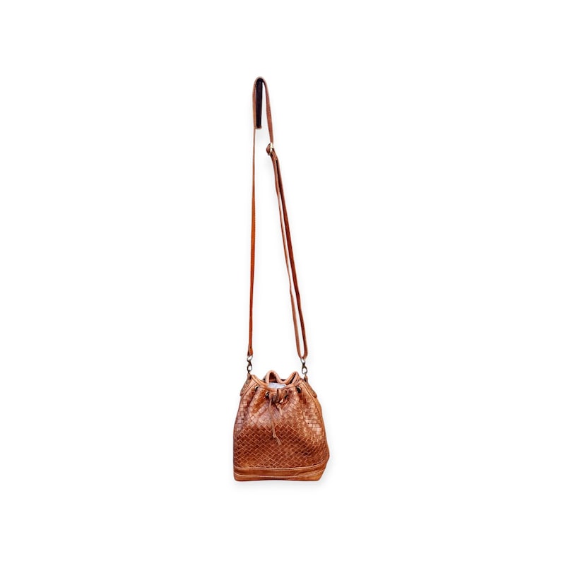 Puede incluir: Un bolso tipo cubo de cuero marr&oacute;n con una textura tejida. El bolso tiene una correa larga y ajustable para llevarlo cruzado. El bolso est&aacute; suspendido sobre un fondo blanco.