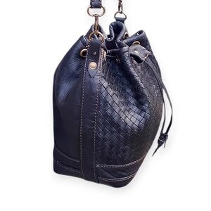 Puede incluir: Bolso tipo cubo de cuero negro con un patr&oacute;n tejido en la mitad inferior y cierre de cord&oacute;n. El bolso tiene una correa para el hombro y herrajes dorados. Las costuras son de un color m&aacute;s claro, contrastando con el cuero oscuro.