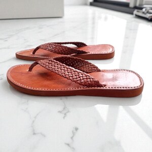 May include: A pair of brown leather flip-flops with woven straps. The sandals have a classic thong design and a stitched sole. The leather appears to be a rich, warm brown colour, and the sandals are displayed on a light-coloured surface.