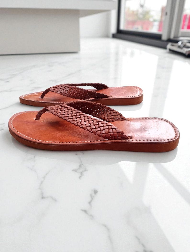 May include: A pair of brown leather flip-flops with woven straps. The sandals have a classic thong design and a stitched sole. The leather appears to be a rich, warm brown colour, and the sandals are displayed on a light-coloured surface.
