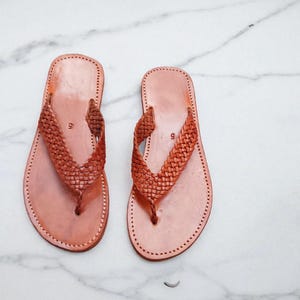 May include: A pair of light brown leather sandals. The sandals feature a woven strap design and stitched soles. The sandals are displayed on a white marble surface. The sandals are suitable for warm weather.