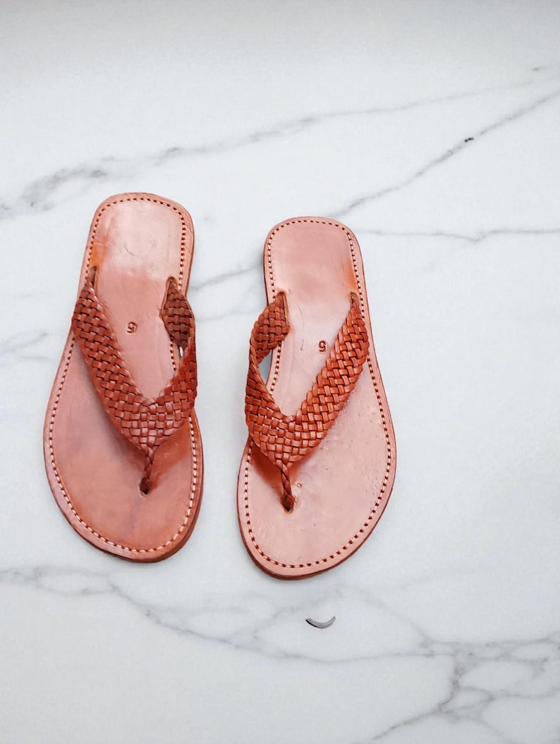 May include: A pair of light brown leather sandals. The sandals feature a woven strap design and stitched soles. The sandals are displayed on a white marble surface. The sandals are suitable for warm weather.