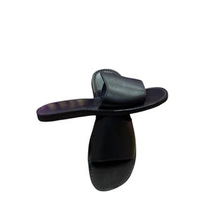 May include: A pair of black leather slide sandals. The sandals have a simple design with a single strap across the top of the foot and a flat sole. The leather appears smooth and the sandals are set against a white background.