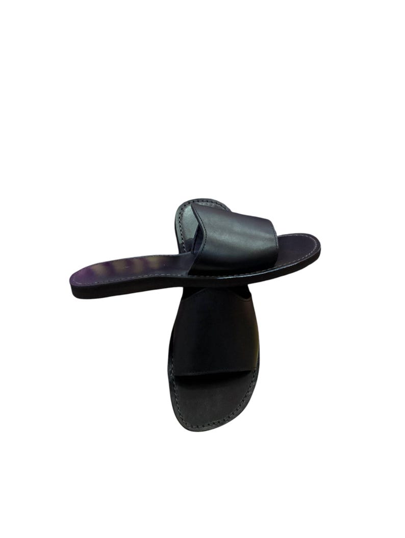 May include: A pair of black leather slide sandals. The sandals have a simple design with a single strap across the top of the foot and a flat sole. The leather appears smooth and the sandals are set against a white background.