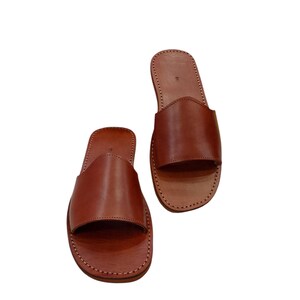 May include: A pair of brown leather slide sandals. The sandals have a simple design with a wide strap across the top of the foot and a stitched sole. The leather appears to be smooth and has a natural finish.