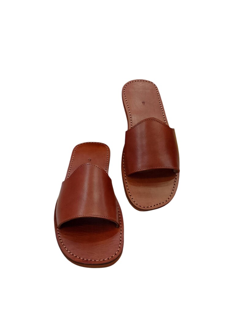 May include: A pair of brown leather slide sandals. The sandals have a simple design with a wide strap across the top of the foot and a stitched sole. The leather appears to be smooth and has a natural finish.