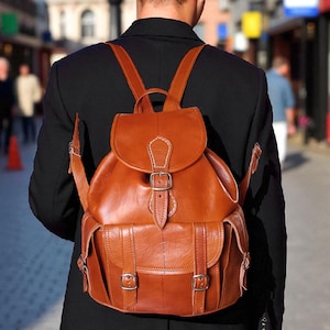 Pode incluir: Uma mochila de couro castanho com fecho de aba e fivela, e dois bolsos frontais. A mochila tem alças ajustáveis e é usada por uma pessoa com um casaco preto, caminhando por uma rua.