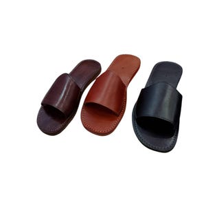 May include: Three leather slide sandals in varying shades of brown and black. The sandals have a simple design with a single strap across the top and are stitched around the edges. The colors include dark brown, medium brown, and black.