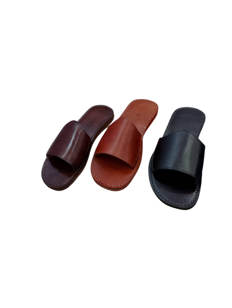 May include: Three leather slide sandals in varying shades of brown and black. The sandals have a simple design with a single strap across the top and are stitched around the edges. The colors include dark brown, medium brown, and black.