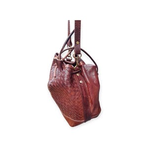 Puede incluir: Un bolso tipo cubo de cuero marr&oacute;n con un patr&oacute;n tejido en la parte delantera y una parte trasera de cuero liso. El bolso tiene un cierre de cord&oacute;n, una correa para el hombro y un asa m&aacute;s peque&ntilde;a. Los herrajes son de color dorado.