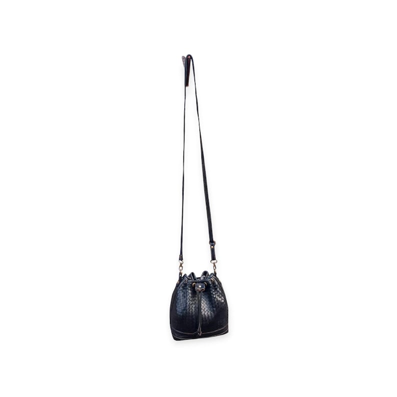 Puede incluir: Un bolso negro con forma de cubo con una textura tejida y una correa larga y ajustable. El bolso tiene un cierre de cord&oacute;n y herrajes dorados. El bolso est&aacute; suspendido sobre un fondo blanco.