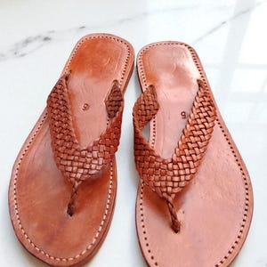 May include: A pair of brown leather sandals. The sandals feature a woven strap design and a thong style. The edges are stitched with white thread. The sandals are displayed on a white surface.