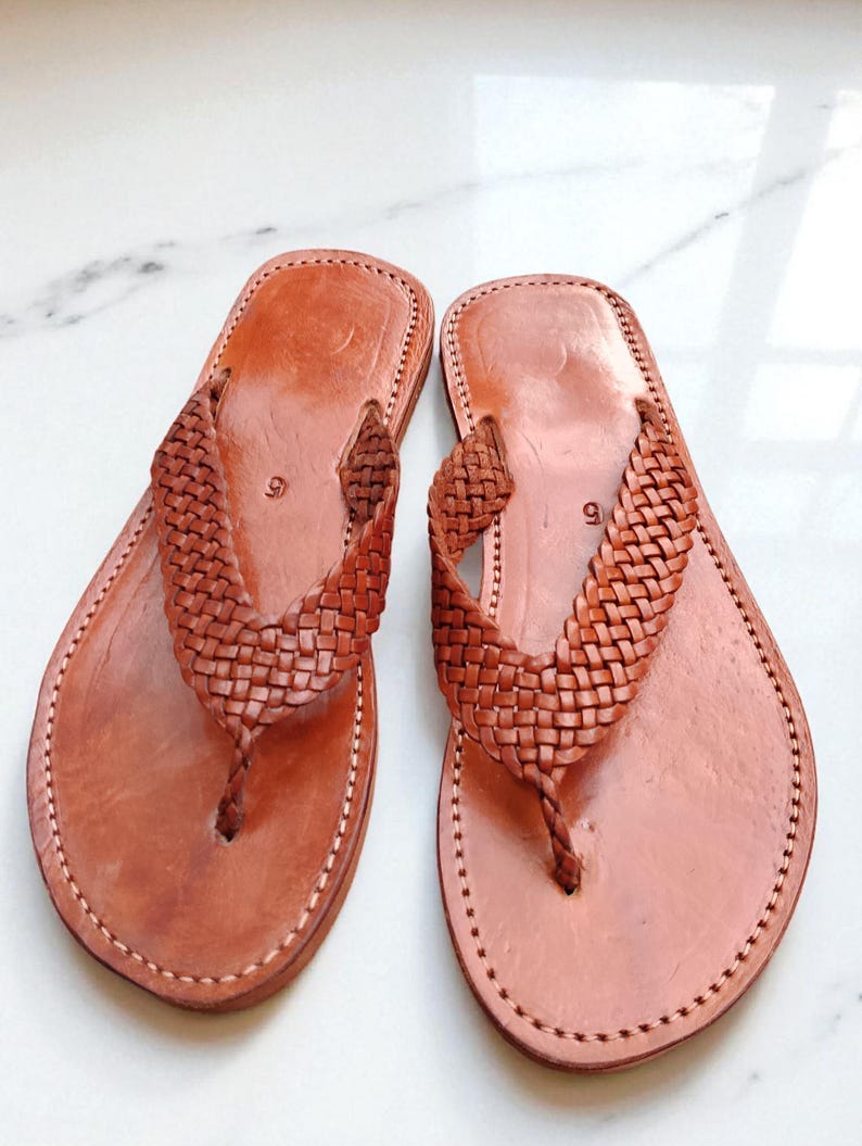 May include: A pair of brown leather sandals. The sandals feature a woven strap design and a thong style. The edges are stitched with white thread. The sandals are displayed on a white surface.
