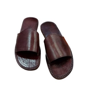 May include: A pair of brown leather slide sandals. The sandals have a simple design with a single strap across the top of the foot and a stitched sole. The leather has a smooth, polished appearance.