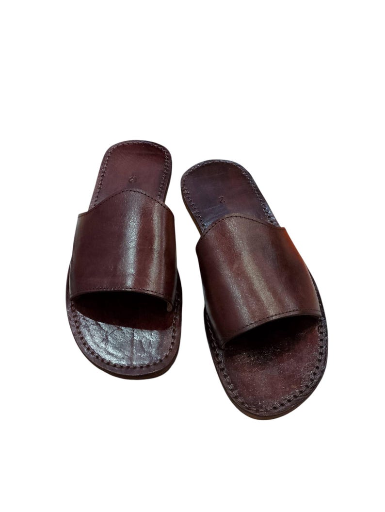 May include: A pair of brown leather slide sandals. The sandals have a simple design with a single strap across the top of the foot and a stitched sole. The leather has a smooth, polished appearance.