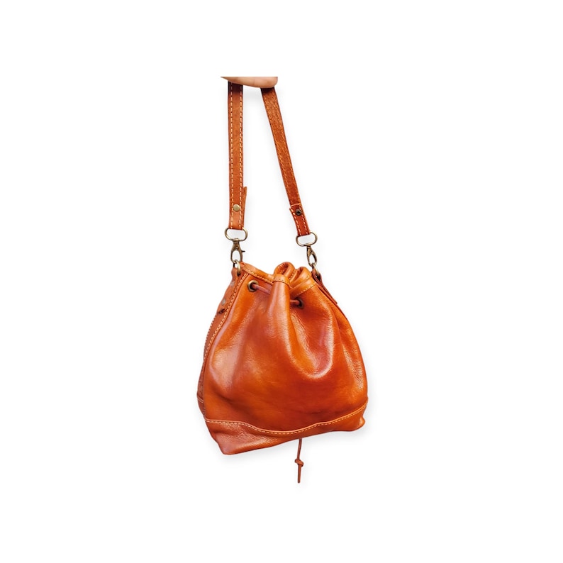 Puede incluir: Un bolso de cuero marr&oacute;n con cierre de cord&oacute;n. El bolso tiene una correa para el hombro y un asa. El cuero es de color marr&oacute;n rojizo c&aacute;lido. El bolso est&aacute; suspendido sobre un fondo blanco.