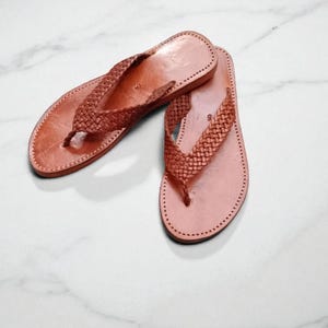 May include: A pair of light brown leather flip-flops. The sandals feature a woven strap design and are stitched around the edges. The flip-flops are displayed on a white marble surface.