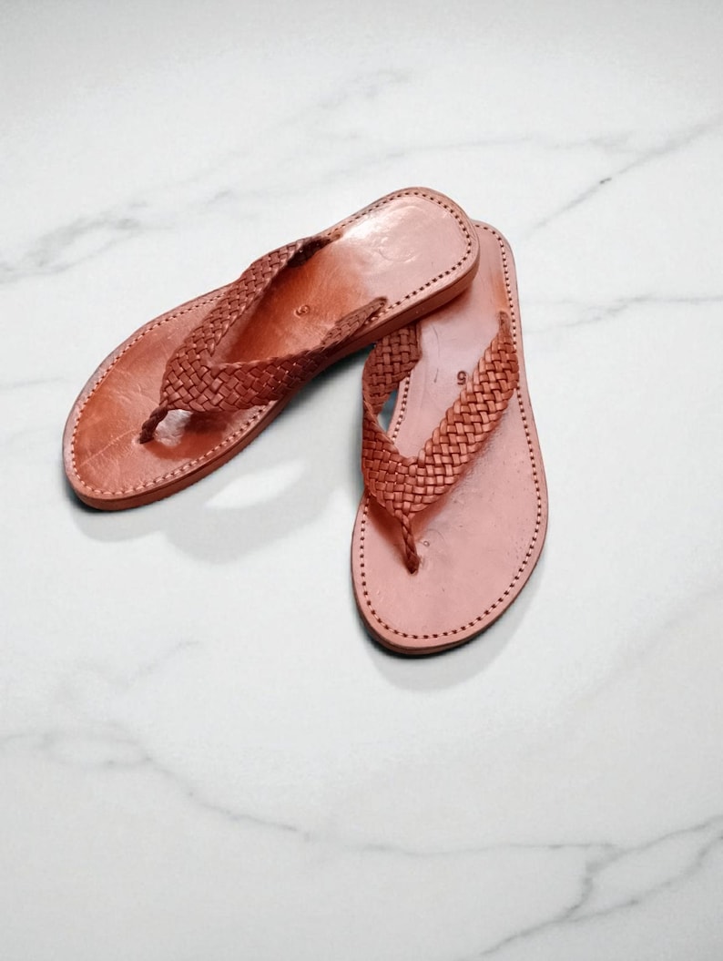 May include: A pair of light brown leather flip-flops. The sandals feature a woven strap design and are stitched around the edges. The flip-flops are displayed on a white marble surface.