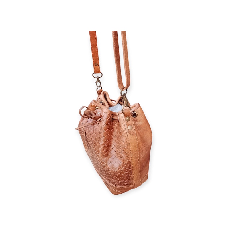 Puede incluir: Bolso tipo cubo de cuero color bronceado con un panel frontal tejido y cierre de cord&oacute;n. El bolso tiene una correa larga y ajustable y herrajes met&aacute;licos. El bolso mide aproximadamente 25 cm de alto y 20 cm de ancho.