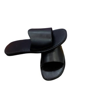 May include: A pair of black leather slide sandals. The sandals have a simple design with a wide strap across the top of the foot and a stitched sole. The sandals are shown on a white background.