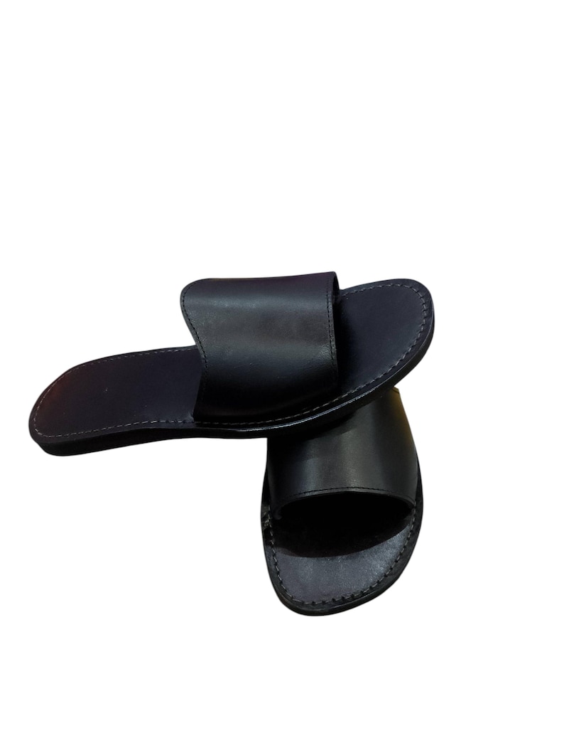 May include: A pair of black leather slide sandals. The sandals have a simple design with a wide strap across the top of the foot and a stitched sole. The sandals are shown on a white background.