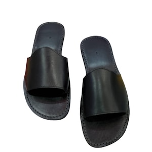 May include: A pair of black leather slide sandals. The sandals have a simple design with a wide strap across the top of the foot and a stitched sole. The sandals are shown on a white background.