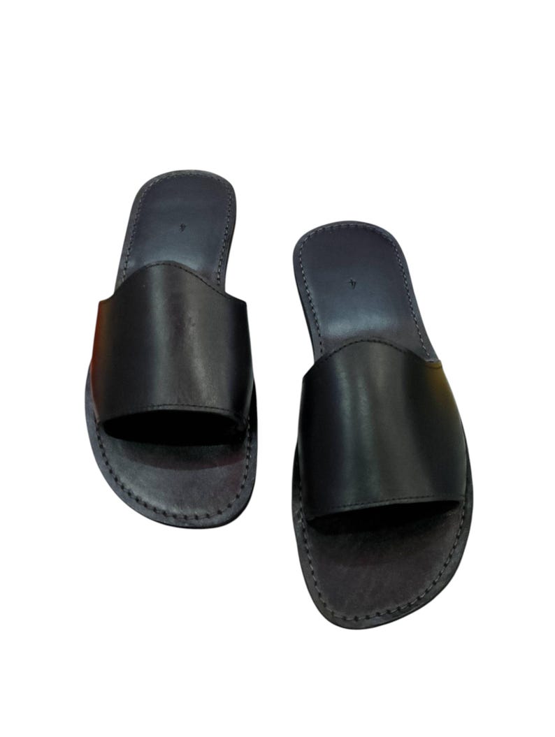 May include: A pair of black leather slide sandals. The sandals have a simple design with a wide strap across the top of the foot and a stitched sole. The sandals are shown on a white background.