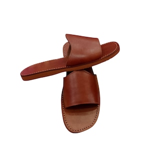 May include: Two brown leather slide sandals. The sandals have a simple design with a wide strap across the top of the foot and a flat sole. The leather appears to be smooth and the sandals are stitched around the edges.