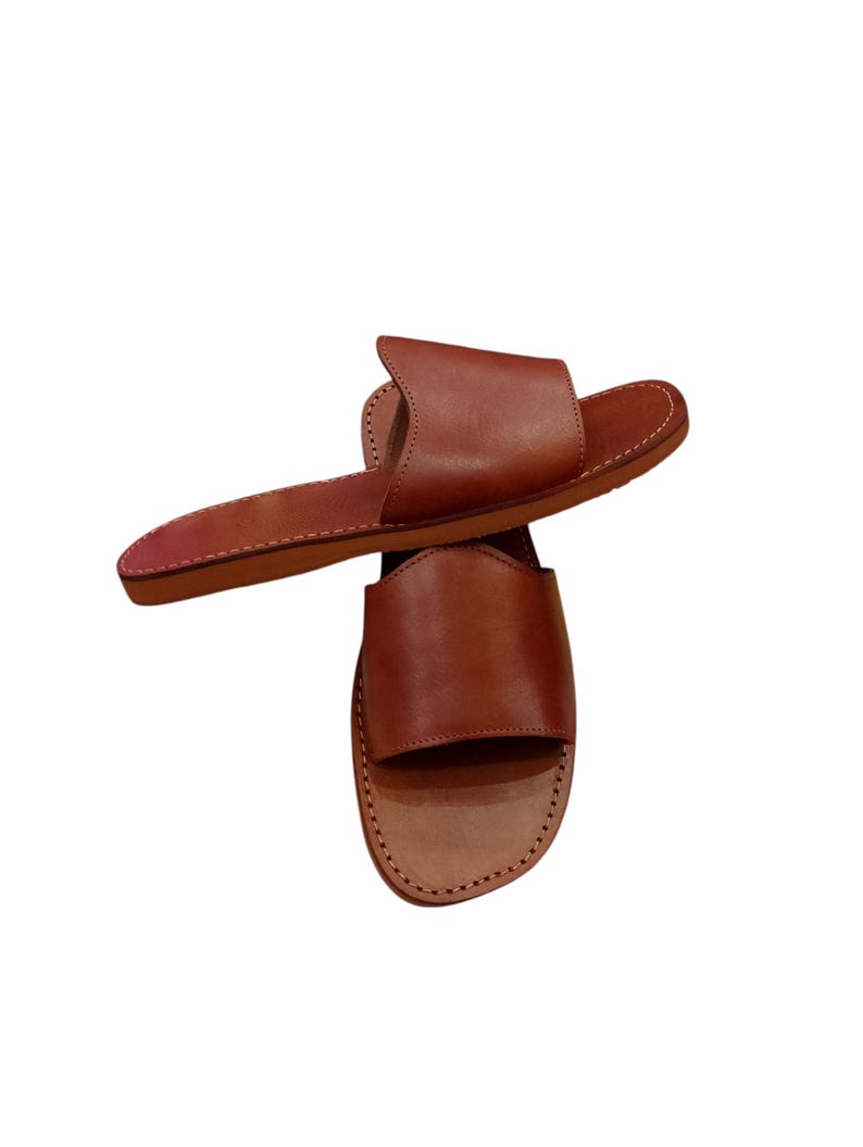 May include: Two brown leather slide sandals. The sandals have a simple design with a wide strap across the top of the foot and a flat sole. The leather appears to be smooth and the sandals are stitched around the edges.