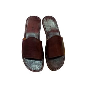 May include: A pair of brown leather sandals. The sandals have a simple design with a wide strap across the top and a stitched sole. The leather appears to be a rich, dark brown color. The sandals are shown on a white background.