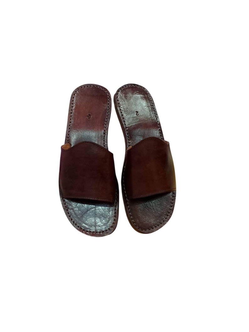 May include: A pair of brown leather sandals. The sandals have a simple design with a wide strap across the top and a stitched sole. The leather appears to be a rich, dark brown color. The sandals are shown on a white background.