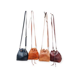 Puede incluir: Cuatro bolsos de cuero tipo cubo en varios tonos de marr&oacute;n y negro cuelgan sobre un fondo blanco. Los bolsos tienen texturas tejidas, cierres de cord&oacute;n y correas largas ajustables. Los colores incluyen negro, naranja, marr&oacute;n claro y burdeos.
