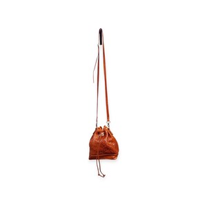 Puede incluir: Un bolso tipo cubo de cuero marr&oacute;n con textura tejida, colgado de una fina correa. El bolso tiene un cierre de cord&oacute;n y una correa de hombro larga y ajustable. El bolso est&aacute; suspendido sobre un fondo blanco.