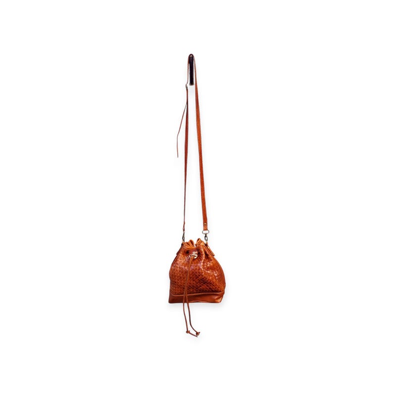 Puede incluir: Un bolso tipo cubo de cuero marr&oacute;n con textura tejida, colgado de una fina correa. El bolso tiene un cierre de cord&oacute;n y una correa de hombro larga y ajustable. El bolso est&aacute; suspendido sobre un fondo blanco.