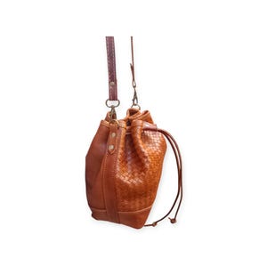 Puede incluir: Bolso tipo cubo de cuero marr&oacute;n con un patr&oacute;n tejido en la parte delantera. Tiene un cierre de cord&oacute;n y una correa larga y ajustable. El bolso est&aacute; hecho de cuero marr&oacute;n c&aacute;lido y tiene un dise&ntilde;o cl&aacute;sico y atemporal.