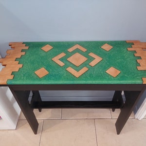 Peut inclure: Une table en bois et verte avec un motif géométrique sur le dessus. La table a une base noire et une bordure en bois autour du dessus.