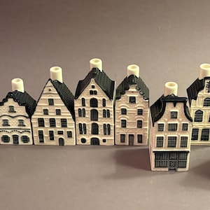 Puede incluir: Una colección de casas de cerámica en miniatura con tejados negros y paredes blancas. Cada casa tiene un diseño arquitectónico único, con ventanas y detalles decorativos. Las casas están dispuestas en fila, creando una exhibición encantadora.