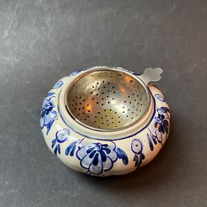 Może przedstawiać: Biały ceramiczny zaparzacz do herbaty z niebiesko-białym wzorem kwiatowym. Zaparzacz ma metalowe sitko z uchwytem.