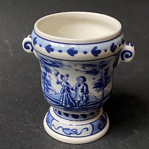 Può includere: Un piccolo vaso in ceramica bianca e blu con due manici. Il vaso presenta una dettagliata illustrazione blu e bianca di due figure in un paesaggio. La base e il bordo sono decorati con motivi blu.