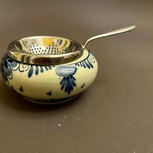 Może przedstawiać: Ceramiczny zaparzacz do herbaty ze stalowym sitkiem i uchwytem. Zaparzacz jest okrągły z kremową podstawą i niebieskimi kwiatowymi wzorami. Uchwyt rozciąga się w prawo, a sitko znajduje się na ceramicznej podstawie.